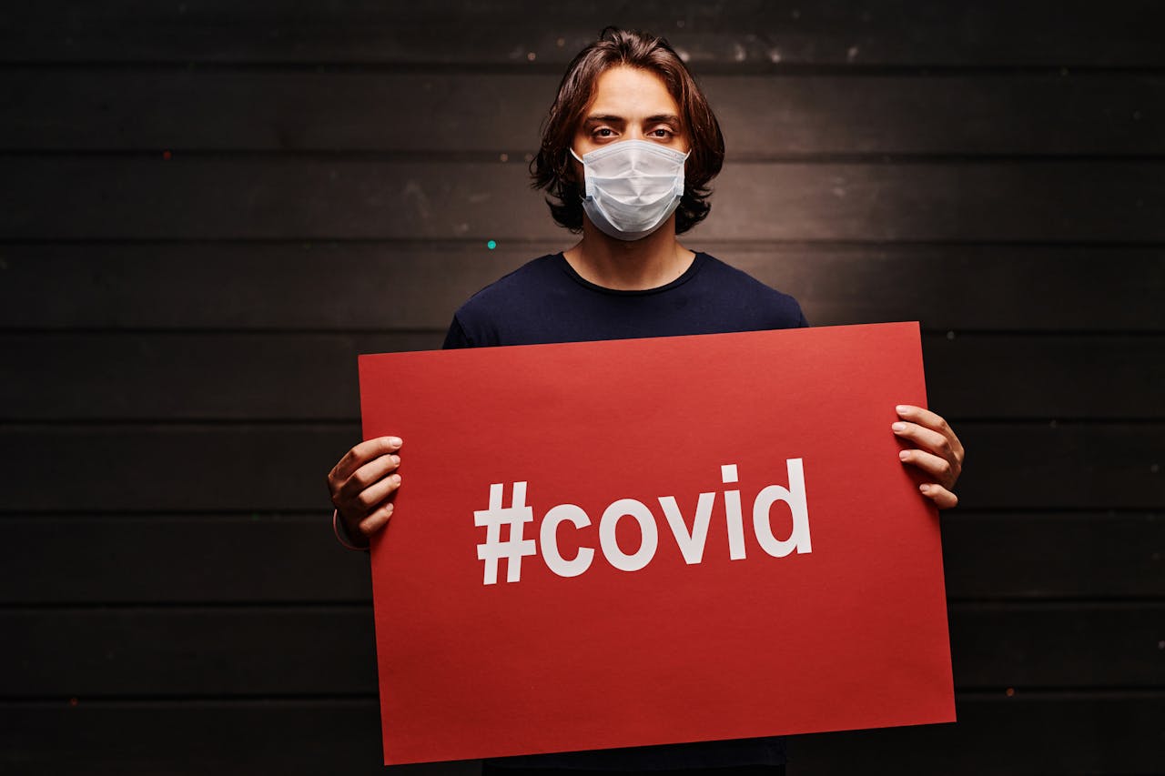 about-01 A man in a face mask holds a red sign with '#covid' indoors. Pandemic awareness.